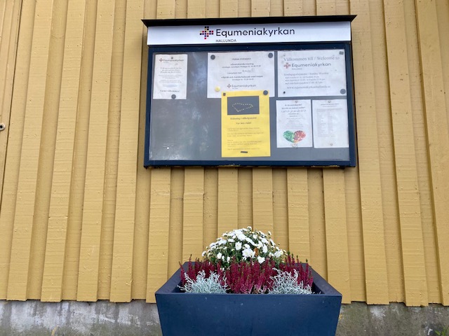 Equmeniakyrkan Hallunda med sin senapsgula träpanel, anslagstavlan med några affischer och framför anslagstavlan på marken en större blåfärhad blomlåda med ljung och andra blommor. Lite av väggen under träpanelen syns också. 